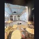 Fotografie zachycuje bicí soupravu umístěnou na pódiu v prostorné sále. Na činelech je vidět nápis Paiste a na blbáku logo REMO. Souprava má průhledn…