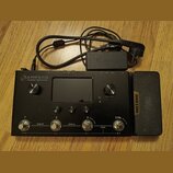 Černý kytarový procesor s nápisem AMPERO Amp Modeler / Effects Processor. Zařízení má displej, dva otočné knoflíky označené VOLUME a MENU / VALUE, tř…