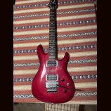 Elektrokytara s červeným lakovaným tělem a hnědým hnedým hmatníkem. Na hlavě je bílé logo Ibanez a na krku nápis RG 5220. Kytara má dva humbuckery, t…