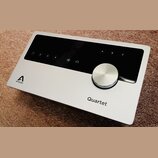 Stříbrný audio rozhraní s černým ovládacím panelem a velkým kovovým otočným knoflíkem. Na šasi je vlevo logo a nápis APOGEE a vpravo nápis Quartet. N…