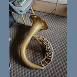 Zlatý mosazný barytonsaxofon položený na koberci. Na zvonu je viditelné kulaté logo, které je však nečitelné. Povrch nástroje vykazuje známky opotřeb…