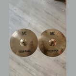 Dvě mosazné činely typu hi-hat položené vedle sebe na světlé podlaze. Oba mají identické nápisy: v horní části „IMC 13" BRILLIANT HI-HAT“ a v dolní č…