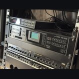 V racku jsou umístěny tři elektronické hudební jednotky. Horní zařízení má nápis YAMAHA a červený digitální displej, prostřední je Casio PROFESSIONAL…