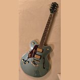 Kytara s polodupatým tělem v modrozelené barvě. Na hlavě a na těle je vidět nápis GRETSCH. Nástroj má dva snímače, červený pickguard a stříbrný hnízd…