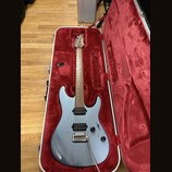 Stříbrná elektrická kytara v otevřeném tvrdém kufru s červeným vnitřkem. Na hlavě hředu je bílý nápis Ibanez. Kytara má dva humbuckery, dva ovládací…