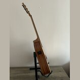 Akustická kytara s přírodním dřevěným povrchem, umístěná na černém stojanu. Na boční straně korpusu je vidět černý vstup pro kabel. Nástroj nemá žádn…