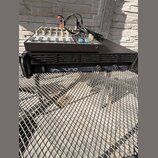 Na černém kovovém mřížkovém povrchu stojí černý rackový zesilovač s nápisem Professional Power Amplifier QSA 1000 a logem Q SOUND. Na zesilovači je u…