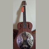 Hnědá akustická kytara typu resonator s kovovým, chromovaným krytem v těle. Na hlavě hředu je bílý nápis Harley Benton a na krku visí oranžová visačk…