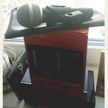 Na černé podložce leží dynamický mikrofon s kovovou mřížkou a černým tělem. Pod ním je červená krabice s nápisy CAM a DR-V1HD. Vedle ní stojí černá k…