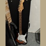 Elektrická kytara s tónem sunburst a bílým pickguardem. Na hlavě hředu je viditelné logo Fender a nápis STRATOCASTER. Kytara má hnědý hmatník a je op…