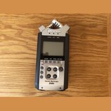 Stříbrno-černý digitální záznamník s nápisy ZOOM v horní části a Handy Recorder H4n ve spodní části. Zařízení má LCD displej, ovládací tlačítka a v h…