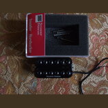 Černý humbucker s dvěma řadami šesti ladících šroubů. Na spodní části je bílý nápis Seymour Duncan. Vedle nástroje je otevřená krabička s červeným št…