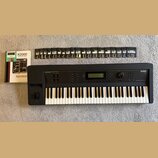 Černý klavír s nápisy KURZWEIL a K2000 V2. Vedle nástroje je položen manuál s názvem K2000 MUSICIAN'S GUIDE a KURZWEIL, spolu s elektronickou deskou.…