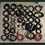 Sada různých audio kabelů a mikrofonů rozložených na dlažbě. Většina kabelů je černá, jsou zde však i bílé a červené varianty s různými typy konektor…