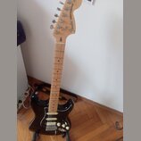 Černá elektrická kytara typu Stratocaster s jasným dřevěným hředníkem a krčkem. Na hlavě je vidět nápis Fender STRATOCASTER. Kytara má tři snímače, b…