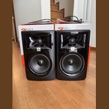 Dva černé regálové reproduktory s lesklým povrchem předními panely. Na obou je umístěno logo JBL. V pozadí jsou dvě bílé krabice s červeným nápisem 3…