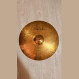 Zlatavý kovový činel s kovaným povrchem. Na horní části je černý nápis Paiste a ve spodní části text Dimensions Hand Hammered Ride. Povrch vykazuje d…