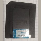 Černá paměťová karta SmartMedia s modrobílou nálepkou. Na nálepce je nápis Smart Media a 128 MB. V horní části těla jsou vyražená písma a čísla: 70NS…