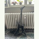 Černá elektrická kytara s ostře hranatým tvarem těla a špičatou hlavou. Na hmatníku jsou vidět bílé polohové značky a na těle jsou dva humbuckery, ov…