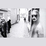 Černobílá fotografie zachycuje muže s dlouhými světlými vlasy a výrazným černým lízením obličeje v ulici s dlážděním. Muž je bez košile a na pravém r…