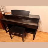 Tmavě hnědý digitální piano v dřevěném provedení s přívodním stolem na noty. Na přední straně je bílý nápis YAMAHA. Součástí je ladící stolek s černý…