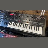 Černý syntezátor s klaviatura značky Roland, model JD-Xi. Horní panel obsahuje červený displej, početné ovládací knofíky a tlačítka rozdělená do sekc…
