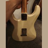 Zadní strana elektrické kytary v krémové barvě. Na kovovém spojovacím plátku u krku je logo Fender a nápis PLAYER PLUS. V dolní části těla je vidět o…