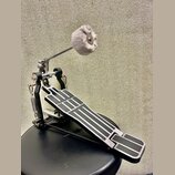 Černý bicí pedál s kovovými prvky a stříbrným stupněm. Na šlapce jsou viditelné bílé vodorovné linky a ornamenty. Beater je vyroben z šedého plstě. N…