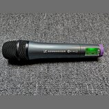 Šedý bezdrátový mikrofon s černou mřížkou a fialovým prstencem u spodní části. Na těle je bílý nápis SENNHEISER s logem a označení ew 100 G2. V zadní…