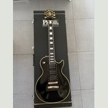 Černá elektrická kytara s bílým lemováním, položená na černém kufru s nápisem PAUL REED SMITH GUITARS. Na hlavě kytary je zlaté logo s nápisem Paul R…