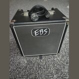 Černý basový kombo zesilovač s kovovými rohy a bílým rámem kolem mřížky. Uprostřed mřížky je oválné logo s nápisem EBS. Na horní straně přístroje lež…