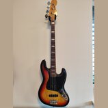 Elektrická basová kytara s tělem v provedení sunburst a černým pickguardem. Na hlavě je nápis JAZZ BASS. Nástroj má hnědý hmatník s bílými polohovými…