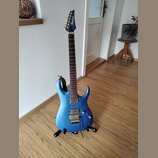 Modrá elektrická kytara s kovovými zlatými komponenty, umístěná na černém stojanu. Na hlavě je viditelné logo Ibanez. Kytara má dva humbuckery, tremo…