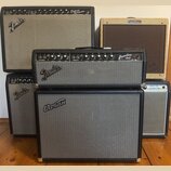 Fotografie zobrazuje čtyři kytarové komba. V horní části jsou dva zesilovače s nápisy Fender Deluxe Reverb Amp a Fender Harvard. Uprostřed stojí komb…