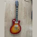 Elektrická kytara s červeným sunburst povrchem a tvarem Les Paul. Na hlavě hředu je bílý nápis Epiphone. Nástroj má dva humbuckery, čtyři otočné knof…