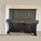 Černý piano v přímém záběru zepředu. Nástroj má zdobný ornament uprostřed klaviaturního víka a tři mosazné pedály. Povrch vykazuje známky opotřebení…