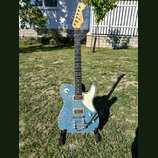 Elektrická kytara s modrým třpytivým povrchem těla a bílým pickguardem. Na hlavě hředu je viditelné logo Fender. Kytara má dva humbuckery, jeden ovlá…