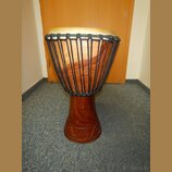Dřevěný buben typu djembe s hnědým povrchem a světlou koženou membránou. Tělo nástroje je v horní části opletené černým šňůrem a v dolní části má vyř…
