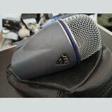 Šedý dynamický mikrofon s kovovou síťovkou a modrým prstenem u hlavy. Na těle je vidět logo v trojúhelníkovém rámečku s nápisem JTS NX 2. Mikrofon le…