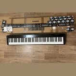 Černý digitální klavír s bílými a černými klávesami. Na horní hraně je nápis CASIO, označení Privia a model PX-S3100 BK. Nástroj leží na dřevěné podl…