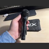 Černý dynamický mikrofon s mřížkou, v držáku. Na těle je bílý nápis AUDIX a pod ním OM-3xb, v dolní části je nápis MADE IN USA a symbol CE. V pozadí…