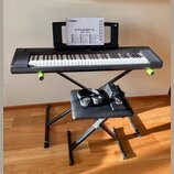 Černý digitální klavír značky YAMAHA umístěný na černém X-stojanu. Na klavíru je nápis YAMAHA a v držáku za ním je manuál s textem piaggero NP-35 NP-…