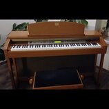Digitální piano v dřevěné barvě s bílými a černými klávesami. Na horní části je nápis Yamaha. Nástroj má ovládací panel s tlačítky a malým displejem,…
