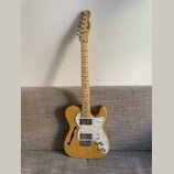 Světle hnědá elektrická kytara s přírodním povrchem dřeva a bílým perleťovým pickguardem. Na hlavě hředu je černý nápis Fender TELECASTER. Tělo kytar…