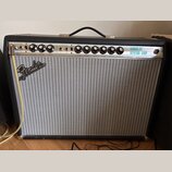 Kytarový kombo zesilovač s černým krytím a stříbrnou síťovkou. Na horním ovládacím panelu jsou nápisy CUSTOM a VINTAGE, značka Fender na přední stran…
