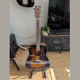 Akustická kytara s tělem v provedení sunburst (černá, hnědá a žlutá). Na hlavě je bílý nápis YAMAHA. Nástroj stojí na černém stojanu v interiéru.