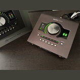 Šedý zvukový interface Apollo Twin značky Universal Audio. Zařízení má velký centrální ovládací knoflík, LED indikátory úrovně a řadu tlačítek s nápi…