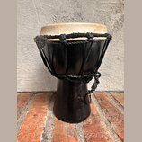 Černý perkusní nástroj typu djembe s béžovou koženou blánou. Tělo je vyrobeno z černého materiálu a horní část je stahována pleteným černým šnůrem. N…