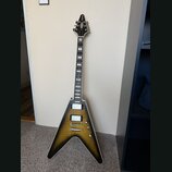 Elektrická kytara s typickým V-tvarem těla v žlutém gradientním provedení s černým okrajem. Na hlavě je viditelné logo Gibson. Kytara má dva humbucke…
