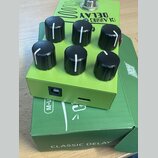 Zelený kytarový efekt s šesti černými oválnými knoflíky. Na horní straně je nápis CLASSIC DELAY a grafický symbol vlnovky. Efekt leží na zelené krabi…
